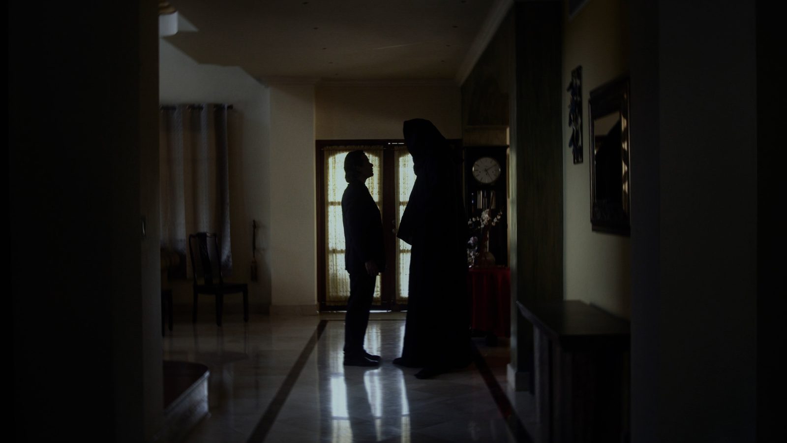 Un pasillo en penumbra con dos figuras conversando al fondo